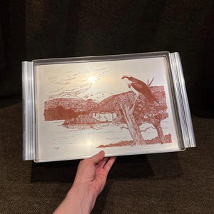 May include: A rectangular silver-toned metal serving tray featuring a copper-coloured etched illustration of an osprey perched on a tree branch overlooking a lake and mountains. The tray has ribbed handles on both sides and a smooth metallic finish.