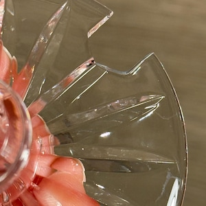 Op de afbeelding: Close-up van een heldere, geslepen glazen serveerschaal met een sterrenpatroon. De schaal heeft een geschulpte rand en wordt gedeeltelijk bedekt door een hand die een roze voorwerp vasthoudt. Het glas heeft een reflecterende kwaliteit.