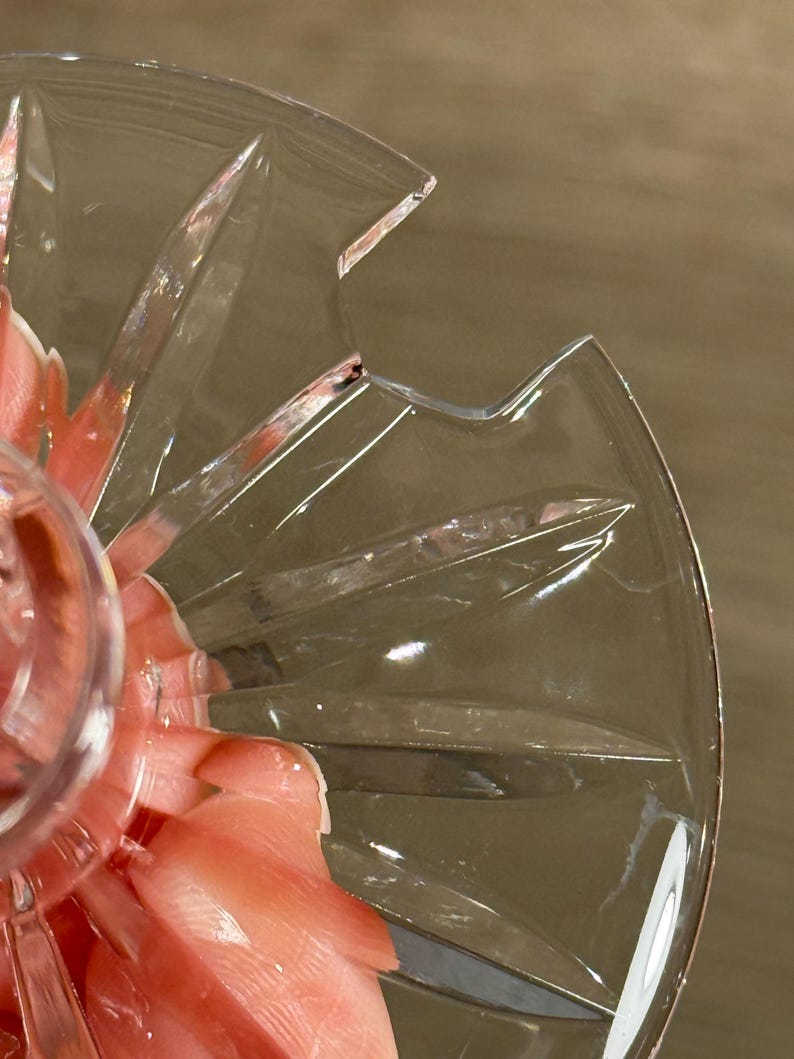 Op de afbeelding: Close-up van een heldere, geslepen glazen serveerschaal met een sterrenpatroon. De schaal heeft een geschulpte rand en wordt gedeeltelijk bedekt door een hand die een roze voorwerp vasthoudt. Het glas heeft een reflecterende kwaliteit.