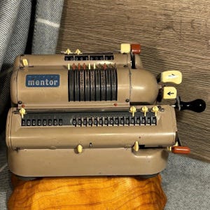 Op de afbeelding: Een vintage grijze metalen Mentor multiplica-calculator met een blauw label en gele en rode knoppen. De calculator heeft een kruk aan de rechterkant en een reeks genummerde wijzerplaten.