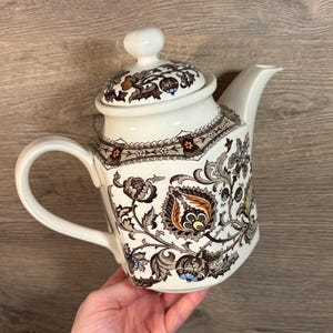 Könnte beinhalten: Eine weiße Teekanne aus Keramik mit einem braunen und goldenen Blumenmuster. Die Teekanne hat einen Deckel und einen Griff.