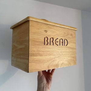 May include: A light oak bread bin with the word "BREAD" cut into the front. The rectangular box has a curved top and is held up by a hand. The wood grain is visible.