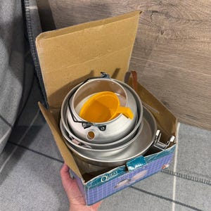 Può includere: Un set di pentole da campeggio in una scatola di cartone. Il set comprende un bollitore argentato, piatti e una tazza di plastica gialla. Sulla scatola è stampata la parola "QUEST". Il set è progettato per cucinare all'aperto.