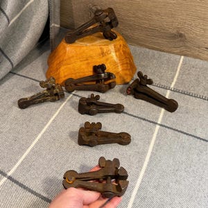 Puede incluir: Una colección de herramientas metálicas antiguas, probablemente para un oficio o comercio específico. Las herramientas son de color marrón oscuro y oxidado, con una variedad de formas y tamaños. Algunas descansan sobre un cuenco de madera, otras sobre una superficie a cuadros gris y blanco.