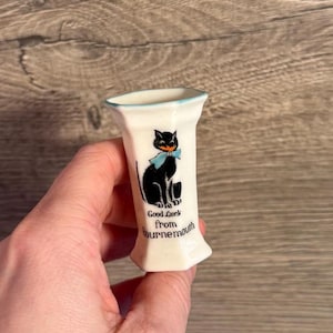 May include: A small, white ceramic vase featuring a black cat illustration with a blue bow tie. The vase has a blue rim and the text "Good Luck from Bournemouth".