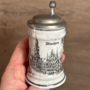 May include: White ceramic beer stein with a silver-coloured metal lid and handle. The stein features a black and white illustration of a city skyline with the word "München" printed above the image.