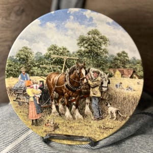 May include: A decorative plate featuring a pastoral scene with two horses pulling a cart. A man and a child are in the foreground, with a dog and a basket of fruit. The background shows a field, trees, and a farmhouse.