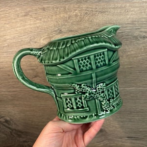 May include: A green ceramic pitcher shaped like a cottage with a thatched roof and a window. The pitcher has a handle and a spout.
