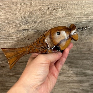 May include: A wooden fish-shaped bottle opener and corkscrew. The opener is carved from light brown wood, with a detailed fish design. The corkscrew is made of silver-colored metal. The fish is held in a hand against a wooden background.