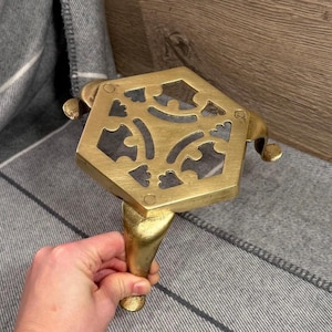 May include: A brass trivet with a hexagonal top featuring decorative cutouts. The trivet has a single leg and two curved handles. The brass has a warm, golden hue, and the trivet is designed to protect surfaces from hot cookware.