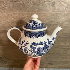 May include: A white and blue ceramic teapot with a lid. The teapot features a classic Willow pattern with blue illustrations of pagodas, birds, and foliage. The handle and spout are white. A decorative piece for tea lovers.
