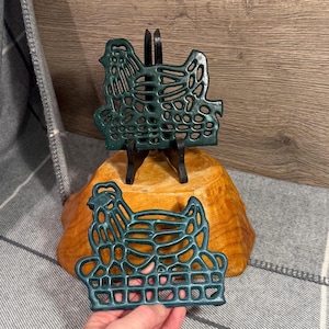 May include: Two teal-colored cast iron chicken trivets. One is displayed on a small black stand, and the other is held in a hand. The trivets have an openwork design and are placed on a wooden surface.