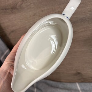 May include: White ceramic gravy boat with a blue and white floral design on the handle. The boat is empty and has a smooth, glossy finish.