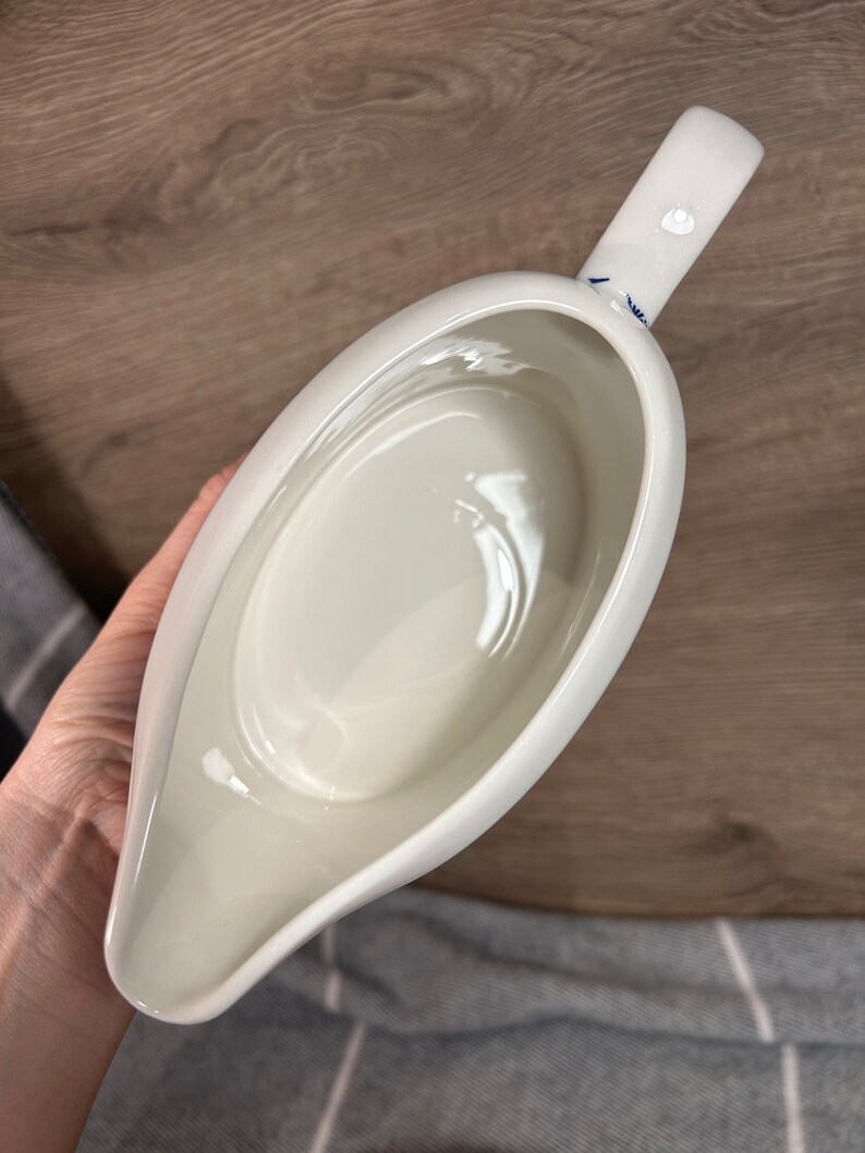 May include: White ceramic gravy boat with a blue and white floral design on the handle. The boat is empty and has a smooth, glossy finish.