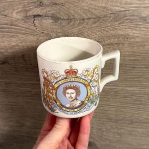 May include: Cream-colored ceramic mug with a gold rim and a square handle. The mug features a portrait of Queen Elizabeth II within a blue oval, surrounded by ornate text and a crown. The text reads "To Commemorate Of The Silver Jubilee Elizabeth II".