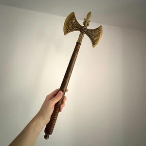 May include: A decorative axe with a wooden handle and a double-bladed head. The head is gold-coloured with intricate dragon designs. The handle is dark brown and has a textured grip. The axe measures approximately 61 cm in length.