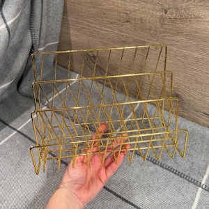 Peut inclure: Un organiseur de bureau en fil métallique doré à trois niveaux. L'organisateur a une forme rectangulaire avec un motif en forme de grille. L'article est tenu par une main, et l'arrière-plan comprend une couverture à carreaux grise et blanche et une surface en bois.