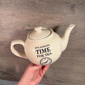Può includere: Una teiera in ceramica color crema con coperchio abbinato, manico curvo e beccuccio. La parte anteriore presenta il testo nero "IT'S ALWAYS TIME FOR TEA" sopra la grafica di un quadrante d'orologio con numeri romani sul corpo arrotondato.