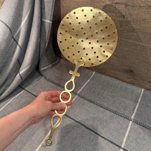 Puede incluir: Un espumadera de cocina de latón vintage con una cabeza circular perforada y un mango adornado. La espumadera se sostiene contra una manta a cuadros gris y blanca y una superficie de madera. La cabeza de la espumadera tiene aproximadamente 15 cm de diámetro.
