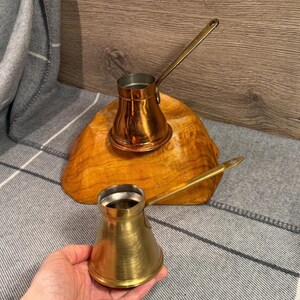 May include: Two small, vintage brass coffee pots with long handles. One pot is resting on a wooden base, while the other is held in a hand. The pots have a classic, conical shape, and the brass has a warm, golden hue.