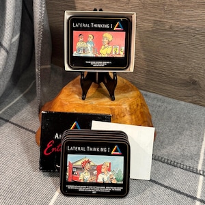 May include: A set of black coasters with the text "Lateral Thinking I" and a colourful graphic. The coasters feature illustrations of people and text. The set is displayed in a white box and on a wooden stand.