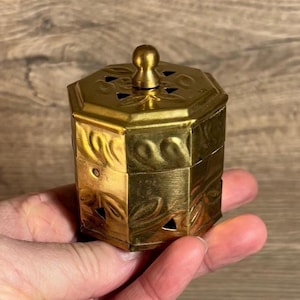 May include: A small, octagonal brass box with a decorative floral pattern and a knob on the lid. The box has small holes around the top and bottom.