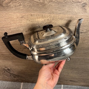 May include: Silver teapot with a black handle and a decorative lid. The teapot has a square body with rounded edges and a polished finish.