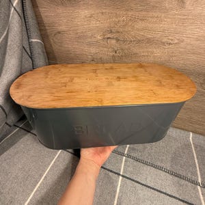 May include: A gray bread box with a bamboo lid. The word "BREAD" is embossed on the front of the box. The lid has the text "George Wilkinson" engraved on it. The bread box is oval-shaped and sits on a gray and white plaid blanket.