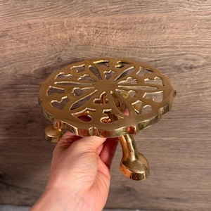 Może przedstawiać: Złoty podkładka pod garnek z dekoracyjnym, ażurowym wzorem. Podkładka ma okrągły kształt z kwiatowym wzorem i trzema zakrzywionymi nóżkami. Wykonana jest z metalu i ma chronić powierzchnie przed gorącymi naczyniami.