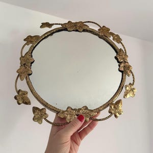 Peut inclure: Un miroir rond avec un cadre doré orné de feuilles et de vignes. Le cadre présente une texture et les feuilles sont finement détaillées. Le miroir est tenu par une main avec du vernis à ongles rouge.