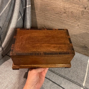 May include: A rectangular wooden box with a hinged lid. The box is a warm brown colour with darker brown accents around the edges. The box is held by a person's hand.
