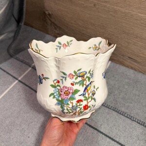 May include: A white ceramic planter with a scalloped rim and gold-tone trim. The exterior features a colourful botanical pattern with pink, red, and blue flowers, green leaves, and a small blue and yellow bird perched on a branch.