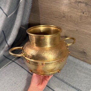 May include: A round, hammered brass vessel featuring a wide opening, two side handles, and a central band accented with small copper-coloured rivets. The metallic surface has a reflective, polished finish with a distinct textured, dimpled pattern.