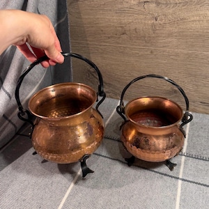 Peut inclure: Deux chaudrons ronds en cuivre avec une texture martelée, chacun doté d'une anse pivotante en métal noir et de trois petits pieds en métal noir. Les chaudrons présentent une finition métallique polie avec des traces d'usure et de patine.