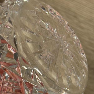 Op de afbeelding: Close-up van een heldere, geslepen glazen kom. De kom heeft een geometrisch patroon met diamant- en sterrenmotieven. Het licht reflecteert op de facetten, waardoor een sprankelend effect ontstaat. De kom is waarschijnlijk bedoeld om te serveren of tentoon te stellen.