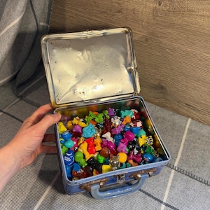 Op de afbeelding: Een vintage blauwe metalen lunchbox gevuld met een verzameling kleurrijk miniatuurspeelgoed. Het speelgoed heeft verschillende vormen en maten, waaronder dieren en abstracte figuren. De lunchbox heeft een handvat en een scharnierend deksel.