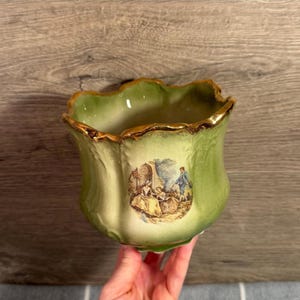 May include: A light green ceramic planter with a scalloped, gold-trimmed rim and a crackled glaze finish. The front features a detailed, circular transfer print depicting two figures in period clothing standing in a garden setting with trees.