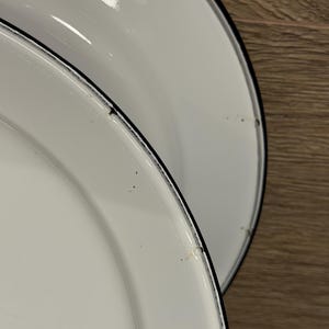 Pode incluir: Dois pratos redondos de esmalte branco com um acabamento liso e brilhante e uma borda preta fina. Os pratos apresentam uma borda ligeiramente elevada e mostram pequenos sinais de desgaste ao longo da borda escura, incluindo pontos.