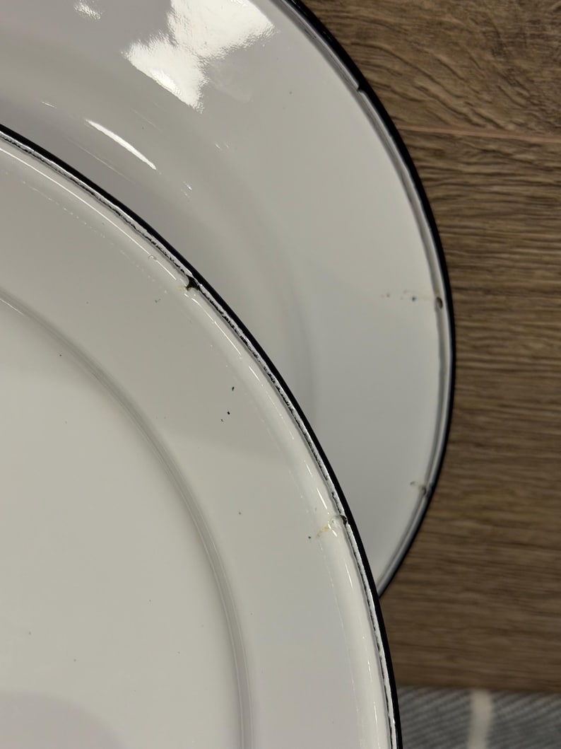 Pode incluir: Dois pratos redondos de esmalte branco com um acabamento liso e brilhante e uma borda preta fina. Os pratos apresentam uma borda ligeiramente elevada e mostram pequenos sinais de desgaste ao longo da borda escura, incluindo pontos.