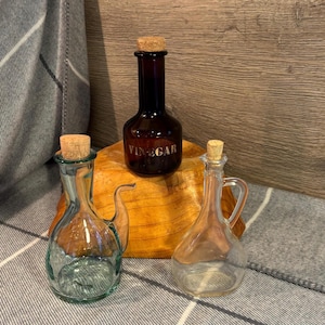 May include: Three glass bottles with cork stoppers. The bottle on the left is green glass with a pour spout. The bottle in the center is brown glass with the word "VINEGAR" etched on the front. The bottle on the right is clear glass with a handle.