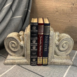 Op de afbeelding: Een paar decoratieve boekenleggers gemaakt van wit geverfd hout met een scroll-ontwerp. De boekenleggers houden drie boeken vast, een met de titel "Oorlog en Vrede" van Tolstoj, een met de titel "Ver weg van de woeste menigte" van Thomas Hardy en een met de titel "Jane Eyre" van Charlotte Brontë.