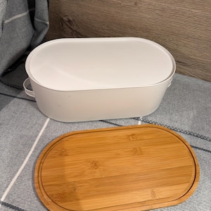 May include: An oval white metal bread box with small side handles and a separate oval bamboo wood lid. The lid features a recessed groove along the perimeter, and the container has a smooth, matte finish with a clean, functional design.