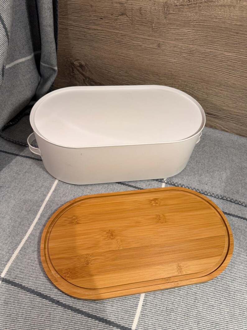 May include: An oval white metal bread box with small side handles and a separate oval bamboo wood lid. The lid features a recessed groove along the perimeter, and the container has a smooth, matte finish with a clean, functional design.