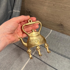 May include: A gold-toned brass wall hook shaped like a tea kettle. The kettle features a central embossed floral design and three downward-curving hooks extending from the base. The top of the kettle includes a handle loop for mounting purposes.