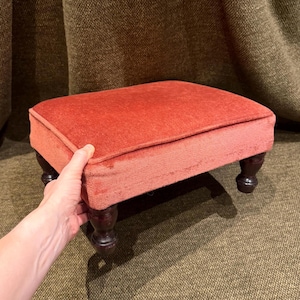 Puede incluir: Un reposapiés rectangular tapizado en una tela texturizada de color naranja quemado con bordes ribeteados. La pieza cuenta con cuatro patas de madera torneada en color marrón oscuro que proporcionan una base estable. El diseño es sencillo.