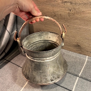 Puede incluir: Un caldero de metal vintage con asa y detalles de cobre. El caldero tiene un cuerpo redondeado y una amplia abertura. El asa está sujeta a los lados con ganchos de cobre. El metal tiene un aspecto envejecido, lo que sugiere edad y uso.