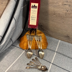 May include: Set of ten silver teaspoons with ornate handles. The spoons are arranged on a wooden surface. A box with the text "Homeland Cutlery, Walker & Co., Sheffield, Improved Nickel Silver" is in the background.