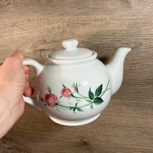 Puede incluir: Tetera de cerámica blanca con tapa y asa. La tetera está decorada con rosas rosas y hojas verdes. La tetera está sujeta por una mano.