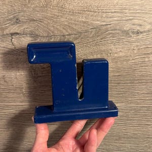 May include: A navy blue metal knife sharpener with a rectangular base and a vertical section with a slot. The sharpener has the word "China" printed on the side.