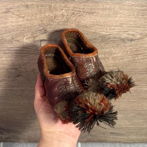 Könnte beinhalten: Ein Paar braune Lederschuhe mit dekorativen Pompons. Die Schuhe haben eine strukturierte Oberfläche und eine kontrastierende, hellbraune Einfassung. Die Pompons sind eine Mischung aus braunen, schwarzen und beigen Fasern.